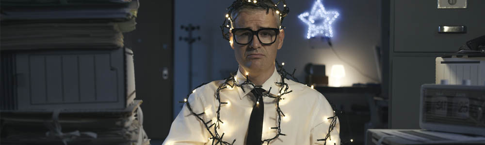 Man in an office wrapped in Christmas lights with work stacked high on his desk, he looks miserable Man in an office wrapped in Christmas lights with work stacked high on his desk, he looks miserable